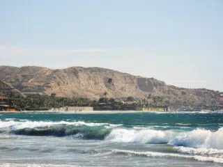 Kitesurfer na falach przy piaszczystej plaży z palmami i wzgórzami-idealne miejsce na wyjazdy kitesurfingowe z KiteAway.