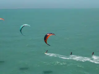 Trzy osoby na wakacjach z kitesurfingiem KiteAway suną po turkusowych falach, kite camp pod niebem Rodos, kolorowe latawce.
