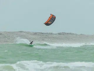 Kitesurfer na zielonych falach przy skalistym brzegu pod chmurami; czarnopomarańczowy latawiec, wyjazdy kitesurfingowe.