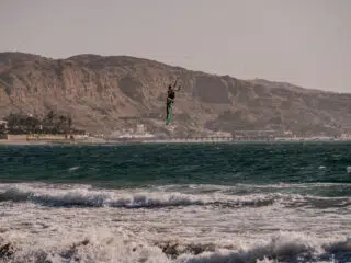 Kitesurfer w powietrzu nad falami przy wybrzeżu z budynkami; idealne miejsce na wyjazdy kitesurfingowe z KiteAway.