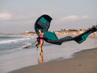 Kobieta w różowym stroju niesie latawiec na plaży podczas wyjazdu kitesurfingowego KiteAway; morze i zabudowa w tle.