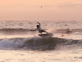 Kitesurfer w białej koszulce płynie o zachodzie na fali podczas wyjazdu KiteAway; w tle czekają inni, wakacje z kitesurfingiem.