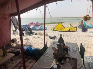 Namiot KiteAway na plaży pełnej sprzętu do kitesurfingu, idealny na wyjazdy kitesurfingowe i kurs kitesurfingu; na tle morza.